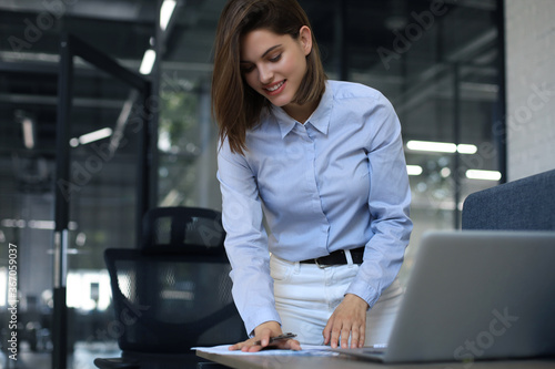 Attractive cheerful business woman checking paper documents in office, working on laptop.