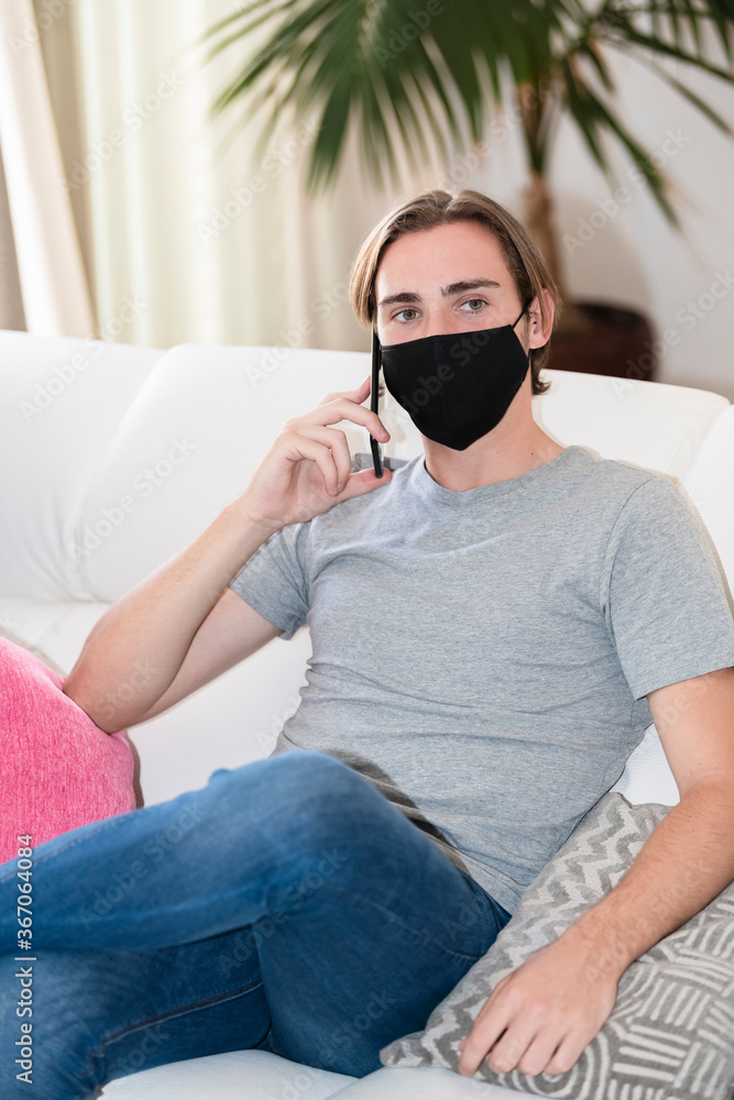 Fototapeta premium young man wearing a face mask sitting on a sofa and talking on his phone. communication concept.