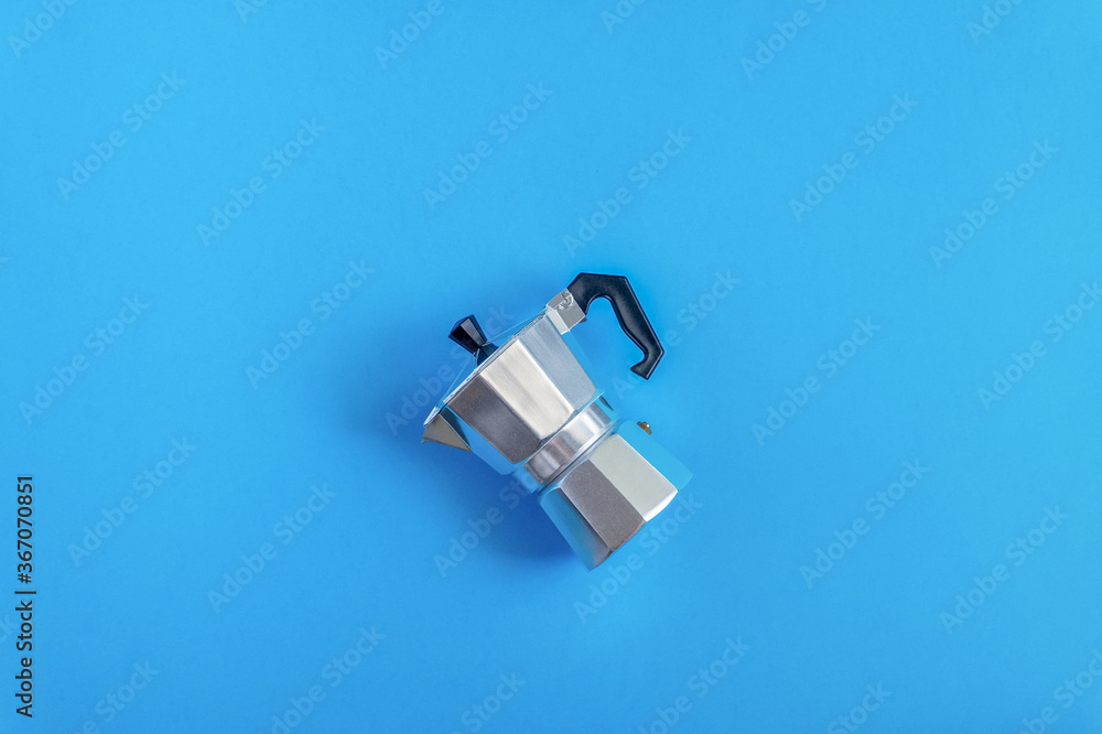 The aluminum geyser coffee maker lies on its side in the center of the blue background. View from above. Horizontal orientation, selective focus. The aluminum geyser coffee maker lies on its side in the center of the blue background. View from above. Horizontal orientation, selective focus.