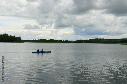 Two people swimming in a kayak or small boat traversing a vast yet shallow river or lake with some dense forest or moor visible in the background and a stormy, moody sky above the horizon