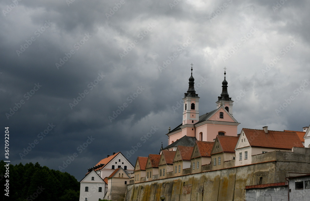 A big christian church or basillica with some concrete walls covering it from all sides seen during a cloudy, stormy day right before a massive tunderstorm on a Polish countryside
