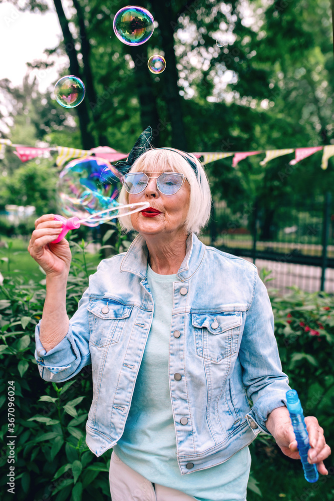 senior (old) stylish woman with gray hair and in blue glasses and jeans jacket blowing bubbles outdoors. Holidays, party, anti age, fun concept senior (old) stylish woman with gray hair and in blue glasses and jeans jacket blowing bubbles outdoors. Holidays, party, anti age, fun concept