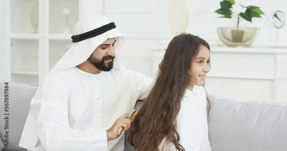 Young handsome Arab man in kandura talking and combing long hair of pretty small teen girl with comb. At home on couch. Muslim father making hair style for little teenage daughter in room. Young handsome Arab man in kandura talking and combing long hair of pretty small teen girl with comb. At home on couch. Muslim father making hair style for little teenage daughter in room.