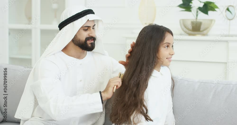 Handsome happy Arabian daddy in dishdasha talking and combing long hair of pretty small teen girl with comb. At home on sofa. Muslim father making hair style for little teenage daughter in living room Handsome happy Arabian daddy in dishdasha talking and combing long hair of pretty small teen girl with comb. At home on sofa. Muslim father making hair style for little teenage daughter in living room