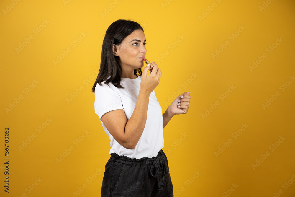 Young beautiful woman painting her lips with a red lipstick. Looking to the left on yellow background. Beauty concept Young beautiful woman painting her lips with a red lipstick. Looking to the left on yellow background. Beauty concept