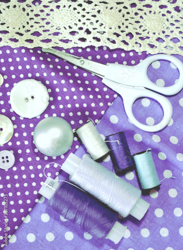 Sewing accessories including white-dotted cotton fabrics, scissors, buttons, sewing spools in lilac and mauve. Scrapbooking and DIY. Hobby and needle work background. 