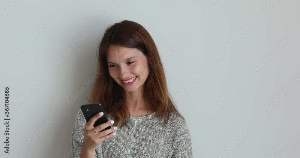 On grey studio background standing woman use mobile phone smile while watching video on internet, social media networks user. Read message feels pleased good news modern tech free time and fun concept On grey studio background standing woman use mobile phone smile while watching video on internet, social media networks user. Read message feels pleased good news modern tech free time and fun concept