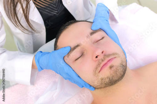 Cosmetologist in gloves is applying cream on client's man face massaging skin, top view. Beautician making beauty facial skincare procedure in clinic, portrait of guy patient. Beauty industry concept.