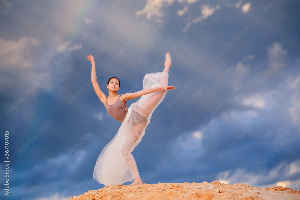 young ballerina in a light long dress stands in an arabesque like a bird, a white skirt develops against the background of the stormy evening sky. A rainbow appears among the clouds.