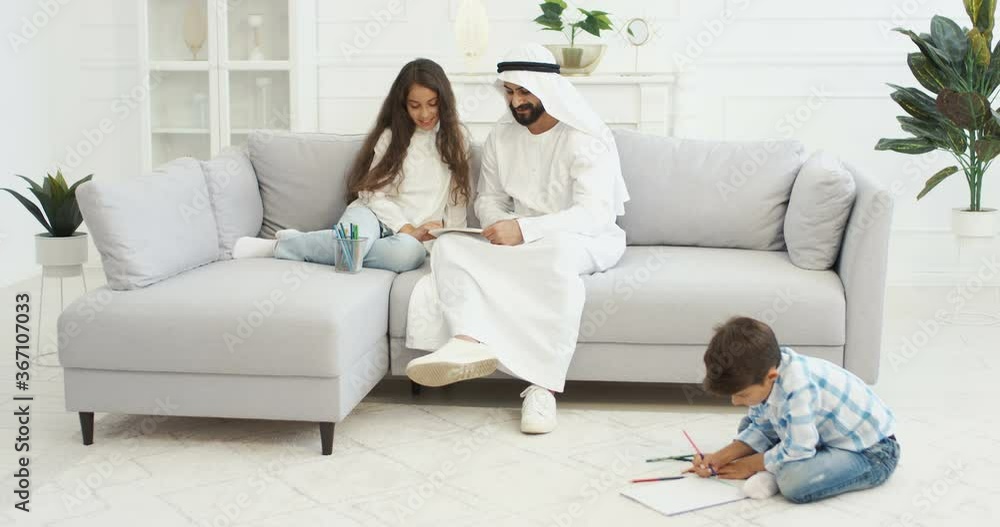 Arab handsome father in dishdasha playing with little children, daughter and son in living room. Muslim dad drawing and coloring pictures. Brother and sister with daddy painting at home and having fun Arab handsome father in dishdasha playing with little children, daughter and son in living room. Muslim dad drawing and coloring pictures. Brother and sister with daddy painting at home and having fun
