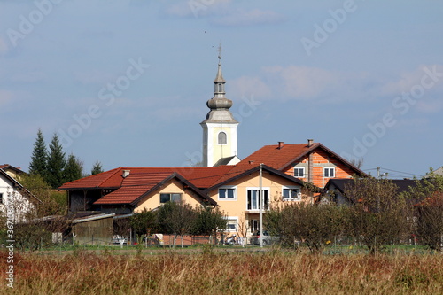 Renovated old church with new facade and metal roof rising above new and unfinished suburban family houses surrounded with dry uncut grass and trees on cloudy blue sky background
