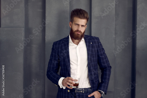 Handsome stylish man in suit stands with glass of whiskey, looks to the camera.