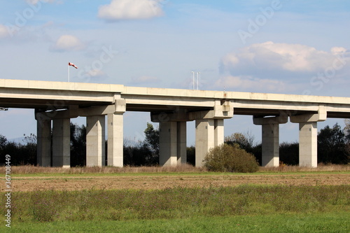 Side view of wide concrete bridge on top of multiple strong thick support columns with dirty old wind direction and speed red and white indicator flag and two white traffic cameras rising high above l
