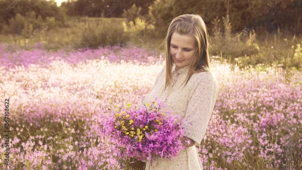 Young woman with a bouquet of wildflowers standing on a sunny blooming meadow with sunset on the background