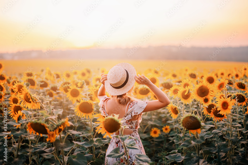 Beautiful summer nature amazing sunny sunset flowering field. Yellow orange sunflower flowers. Young modern woman turned away, enjoys harmony nature, rural relaxation. Girl touches straw hat on head - obrazy, fototapety, plakaty 