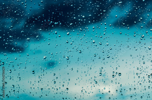 blue rain water drops on a window glass close up with deep dark storm clouds on thr backdrop, colored drop background macro in a blue light