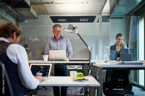 Smiling business colleagues using laptops while working at creative office with glass shields during pandemic