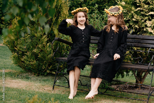 Two little girls in black dresses with wreaths on their heads are sitting on a bench in a dror. Relax in the fresh air and have fun. The concept of children's style and fashion. photo