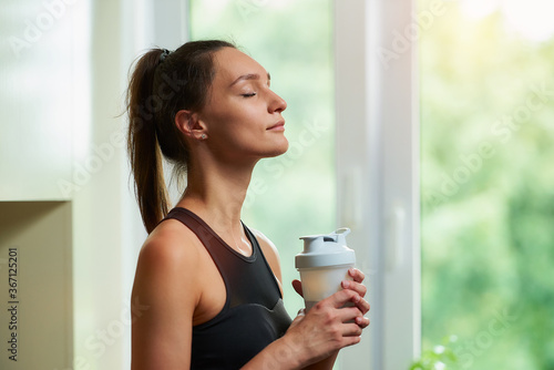 A side view photo of a sporty slim girl with closed eyes in a black workout tight suit is holding the white plastic shaker before training near the window at home.