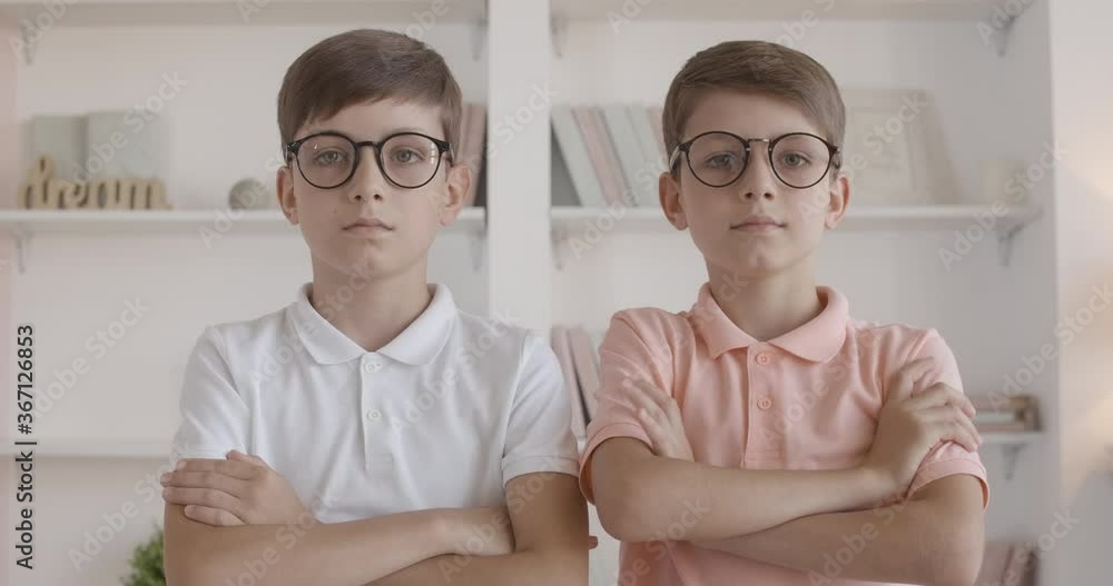 Twin brothers adjusting eyeglasses and crossing hands. Portrait of little Caucasian children posing at home. Smart kids looking at camera with serious facial expression. Twin brothers adjusting eyeglasses and crossing hands. Portrait of little Caucasian children posing at home. Smart kids looking at camera with serious facial expression.