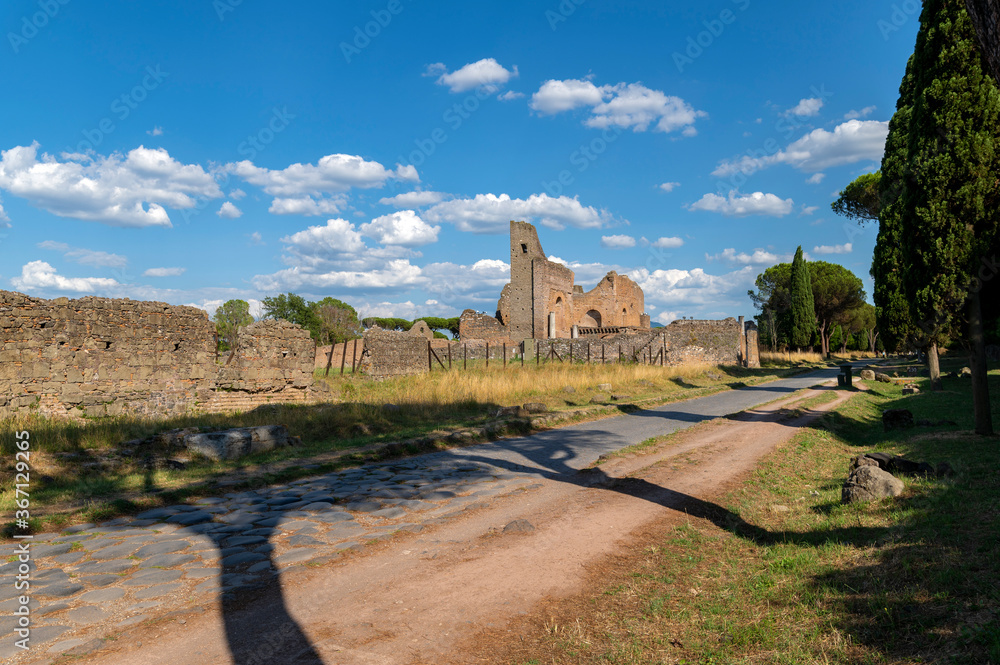 Via Appia Antica, in summer between clouds, blue sky and the shadows of the trees. Next to the ruins of Villa dei Quintili. This road connected Rome to Brindisi. Italy. Maritime pines and cypresses