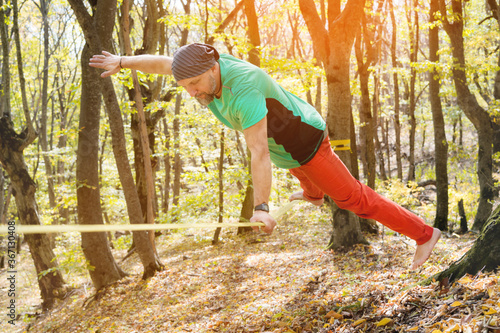 A bearded man of age does a trick on his arm balancing on a taut slackline in the autumn forest. The concept of an active lifestyle for middle-aged people