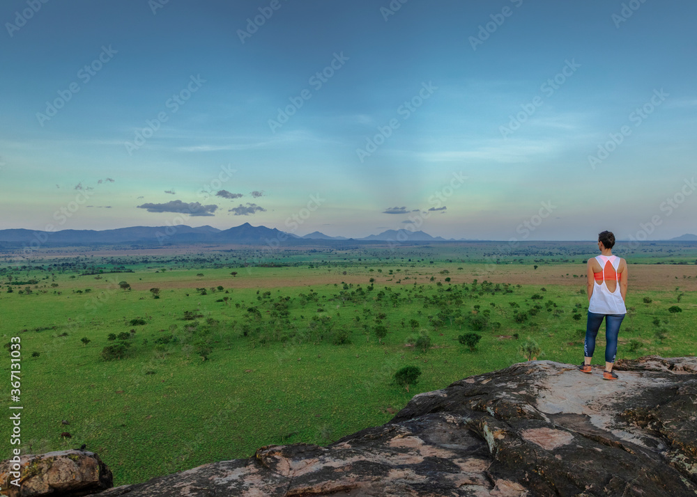 Young woman looking into the horizon of the beautiful landscapes of Kidepo Valley National Park, Uganda, Africa. The woman rests on top of a kopje after a nice hike.