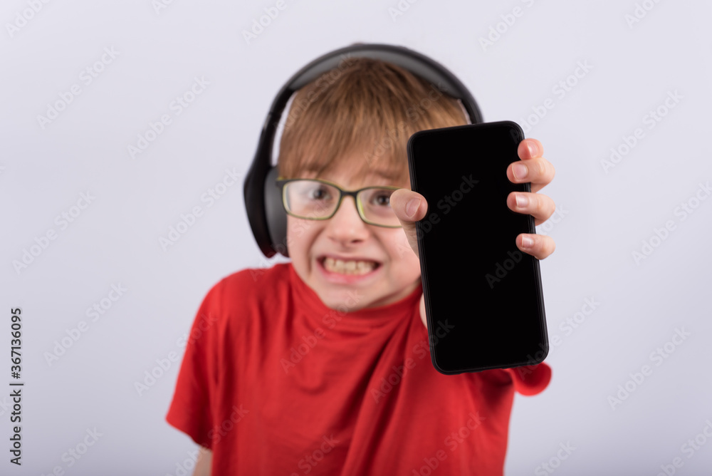 Scared little boy with headphones holding smartphone with blank black screen. Child broke phone. Copy space. Mockup. Scared little boy with headphones holding smartphone with blank black screen. Child broke phone. Copy space. Mockup.
