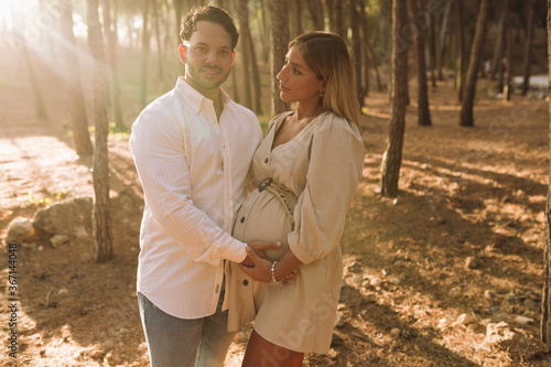 Pregnant couple touching their bellie in a forest and looking at the camera. Blonde pregnant woman with her brunette boyfriend. Blonde pregnant woman in a beige dress. photo