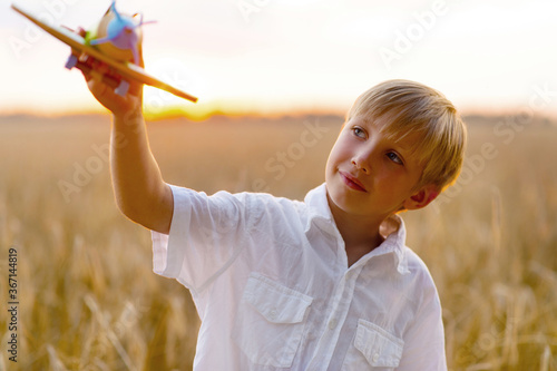 Happy child playing with a toy plane in nature during summer sunset. Boy in a white shirt with a plane in hands on wheat field. Kid holds a wooden airplane and dreams of being a pilot, on the nature