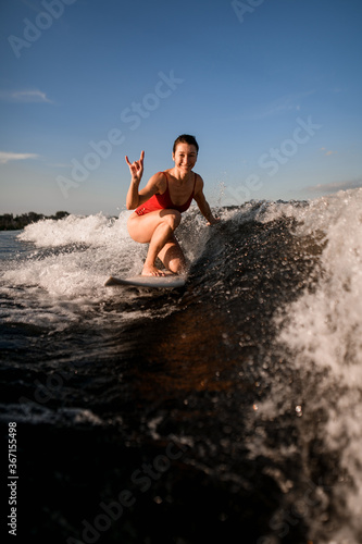 Fototapeta Naklejka Na Ścianę i Meble -  smiling woman rides on surfboard and shows hand gesture