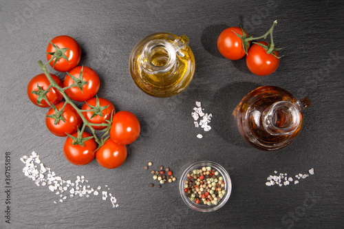 A twig of cherry tomatoes with some coarse salt grains, an oil and a vinegar jug and colorful pepper on a slate slab.