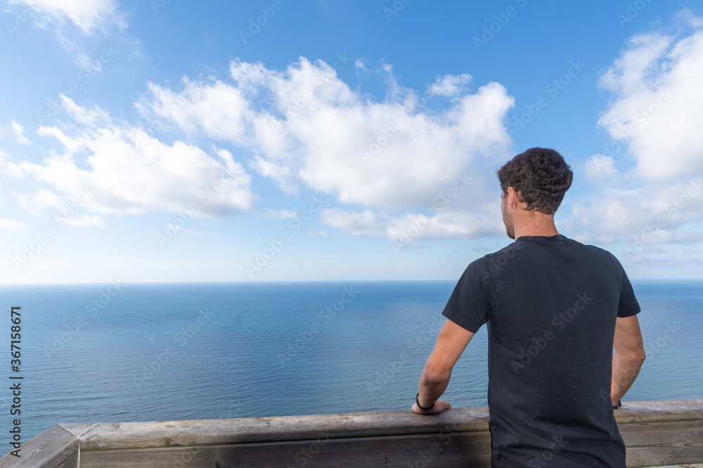 Young man on his back leaning against a wooden railing, looking at a sunny seascape with white clouds, horizontally, with copy space