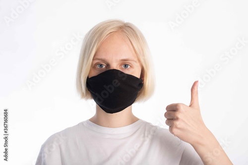 portrait of a young girl with light hair and blue eyes in a black mask (virus deterrent). the girl shows the like sign with her left hand