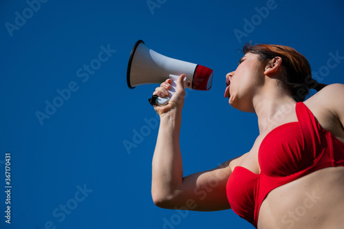 Caucasian woman in red bikini works as a lifeguard on the beach and shouts through a megaphone against a blue sky. A girl in a swimsuit is standing with a loudspeaker. Summer vacation. © Михаил Решетников