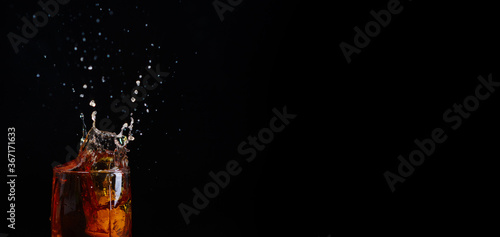 A glass of whiskey with an ice cube falling into it and flying splashes on a black background. Panoramic stretched image