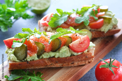 Sandwiches with salmon, avocado, pickled cucumbers, cherry tomatoes and arugula on wooden cutting board