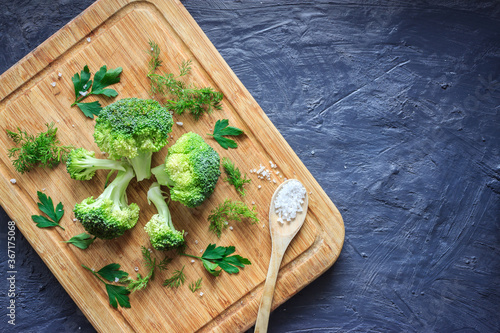 Top view of fresh broccoli, parsley, dill and wooden spoon with salt on grey dark rustic background with copy space. Fresh green vegetables on wooden chopping board. Healthy food concept.