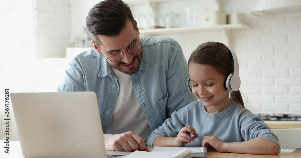 Caring young father helping little primary school learner daughter doing homework, e-learning indoors. Smiling parent explaining education program to small girl in headphones, online study concept. Caring young father helping little primary school learner daughter doing homework, e-learning indoors. Smiling parent explaining education program to small girl in headphones, online study concept.