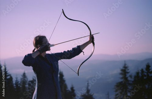 Woman shooting with a bow in the mountains. Young Caucasian female archer shooting with a bow at sunset. Scanned film.