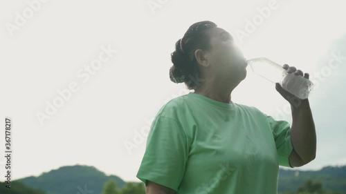 Asian elder female drinking water from plastic bottle after work out at the park, refreshing clean air relaxing after exercising, retied pensioner, human health wellness vitality, sun flare reflection photo