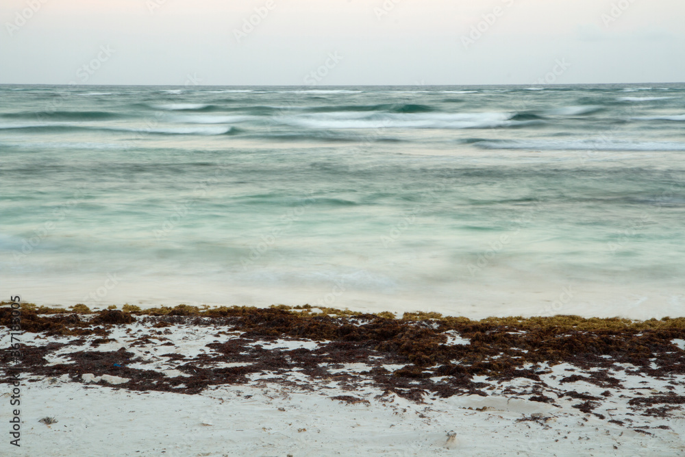 Long exposure. Summer. The beach and turquoise water ocean at sunrise.  The white sand shore with sargassum seaweed and blurred sea waves with a magical dawn light.