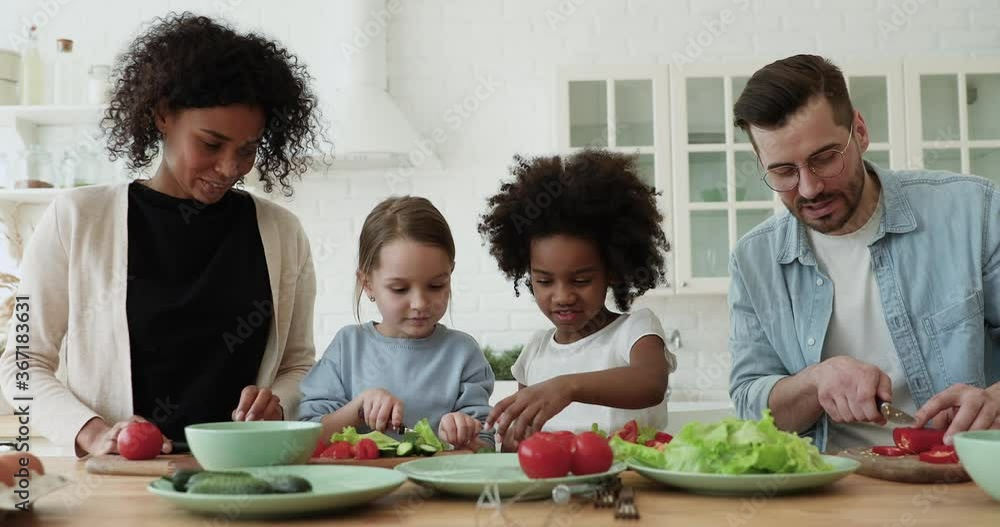 Happy interracial couple teaching little mixed race daughters preparing fresh healthy food in modern kitchen. Smiling multiracial family involved in cooking with adorable small diverse children. Happy interracial couple teaching little mixed race daughters preparing fresh healthy food in modern kitchen. Smiling multiracial family involved in cooking with adorable small diverse children.