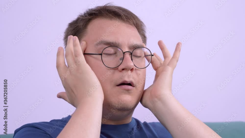 Irritated tired young man sighs nervously and gently rubs temples with hands, takes off glasses, massages the bridge of his nose from fatigue. Irritated tired young man sighs nervously and gently rubs temples with hands, takes off glasses, massages the bridge of his nose from fatigue.