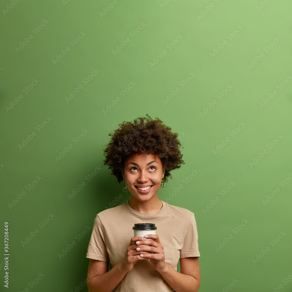 Coffee is never enough. Smiling pleased young woman looks above on empty space, holds hot drink in disposable cup, enjoys caffeine beverage, isolated over green background, notices something up Coffee is never enough. Smiling pleased young woman looks above on empty space, holds hot drink in disposable cup, enjoys caffeine beverage, isolated over green background, notices something up