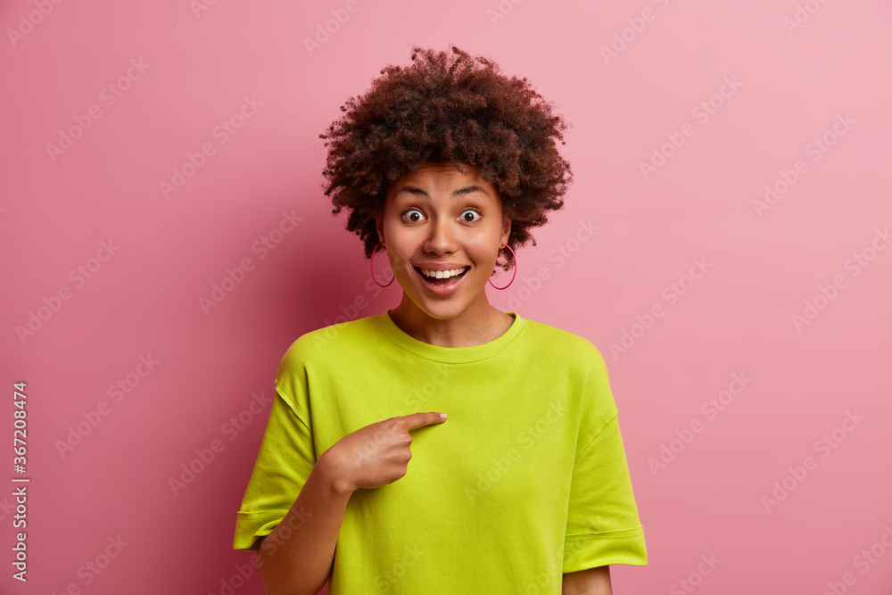 Positive young woman with curly hair points at herself, cannot believe being chosen, picked in someone team, has glad face expression, wears casual green t shirt, isolated over rosy background
