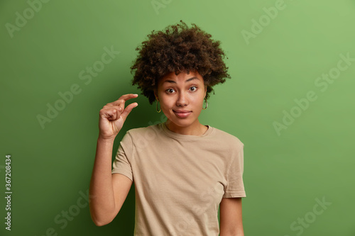 Unimpressed curly woman shows some more or little bit gesture, demonstrates few inches or sentimeter, small amount or low scale sign, dresseed in casual clothes, isolated on green studio wall