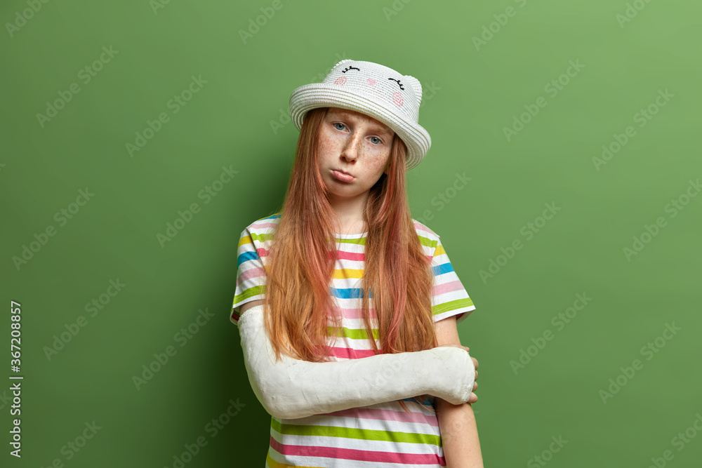 Displeased offended redhead girl with freckled face, being in bad mood after getting trauma, tilts head and purses lips, wears hat and striped t shirt, poses against green studio background. Displeased offended redhead girl with freckled face, being in bad mood after getting trauma, tilts head and purses lips, wears hat and striped t shirt, poses against green studio background.