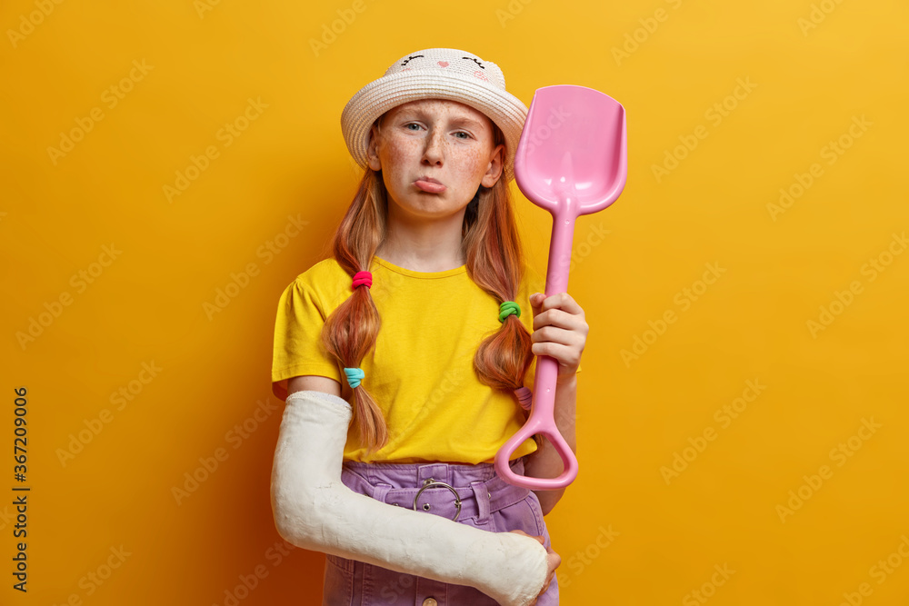 Photo of displeased redhead girl with freckles, purses lips and looks unhappily at camera, cannot play with friends, holds toy shovel, has broken arm in plaster cast, wears hat, t shirt and skirt Photo of displeased redhead girl with freckles, purses lips and looks unhappily at camera, cannot play with friends, holds toy shovel, has broken arm in plaster cast, wears hat, t shirt and skirt