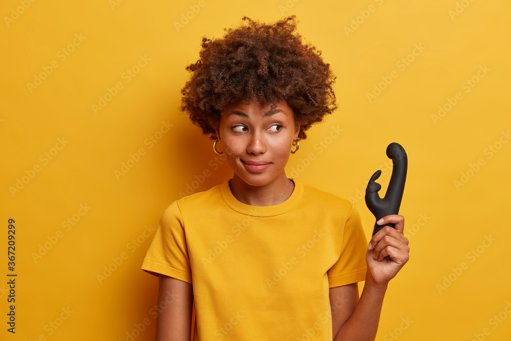 Photo of dark skinned female gives sex tips, recommends buying vibrator used to provide clitoral orgasms and stimulation, enjoys solo masturbation session, gets pleasure isolated on yellow background. Photo of dark skinned female gives sex tips, recommends buying vibrator used to provide clitoral orgasms and stimulation, enjoys solo masturbation session, gets pleasure isolated on yellow background.